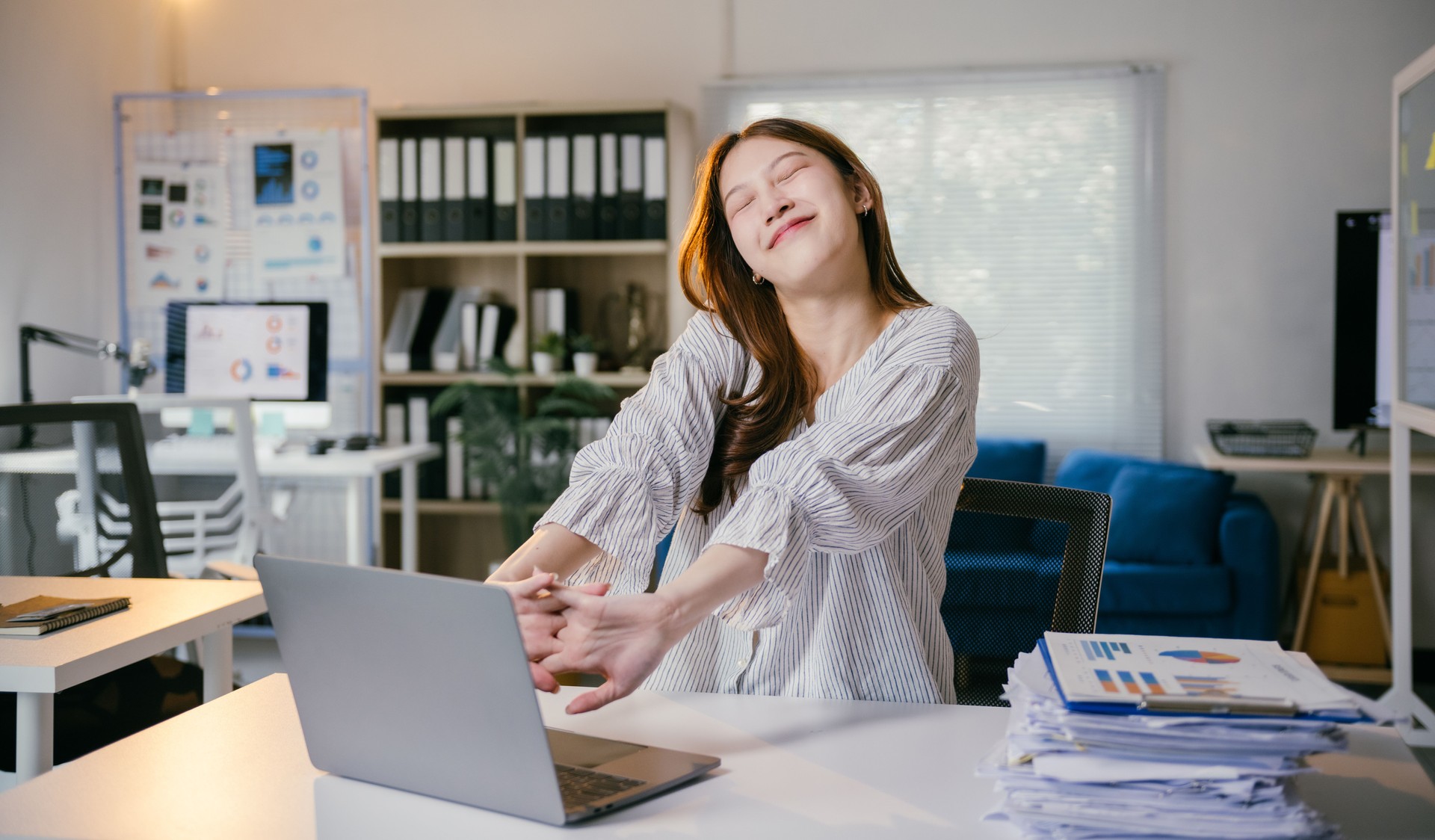 Young happy asian businesswoman stretching arms at workplace after work on laptop computer in modern office, feeling tired after long working day